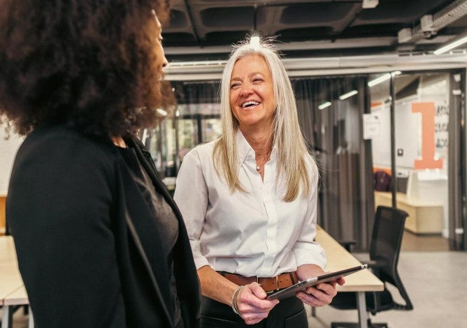 A smiling woman business leader holds a tablet while talking to her female employee in their modern workspace