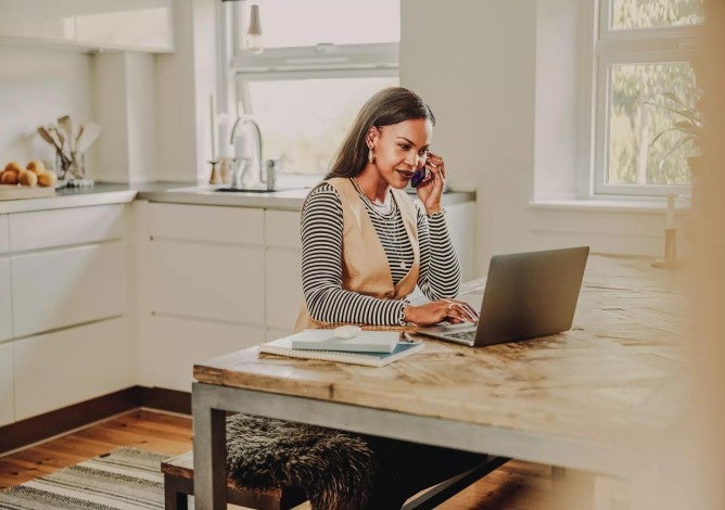 Woman at home working on computer and talking on cellphone.