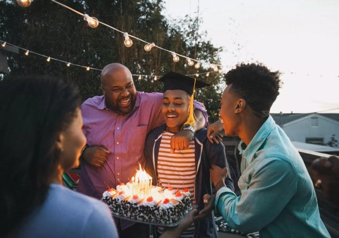 Young man in graduation cap and gown blowing out candles on a cake surrounded by smiling family and friends.