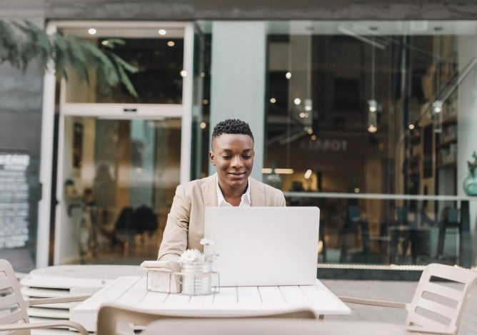 Young man working on his laptop at a patio table outside of a business. 