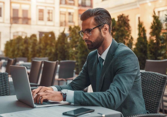 Portrait of bearded businessman in eyeglasses working with laptop while sitting in restaurant outdoors.