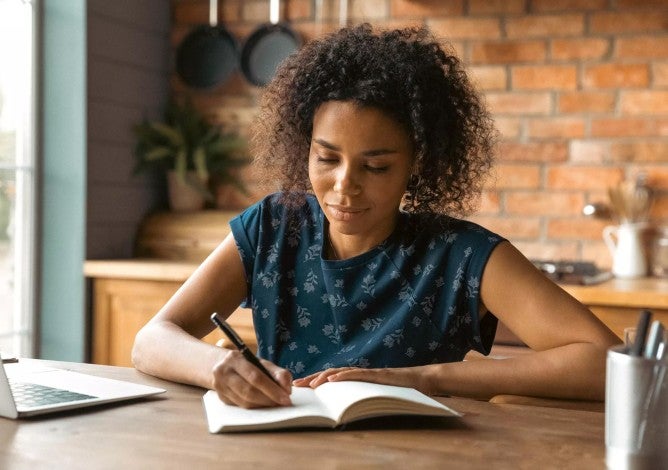 Woman confidently making notes in a notebook while working on her laptop