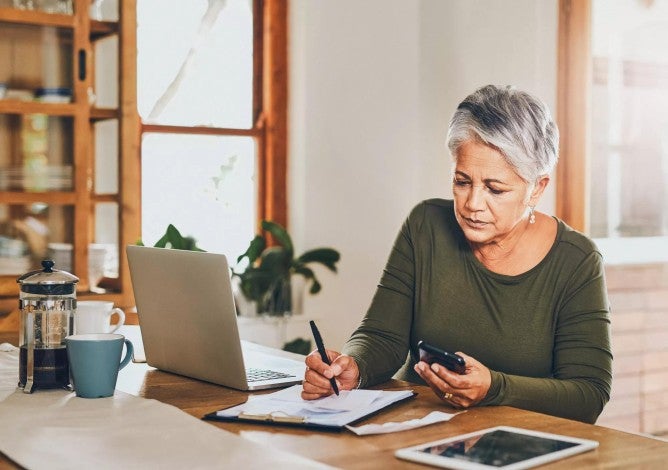 Women working at a table with her laptop and cell phone, writing on paper. 