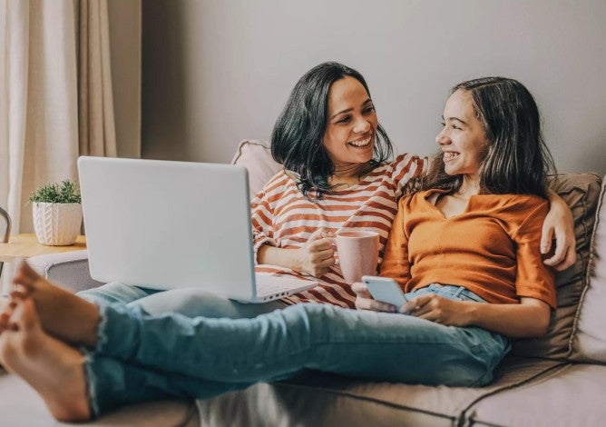 Mother and daughter sitting together on a couch looking at a computer and talking about money.