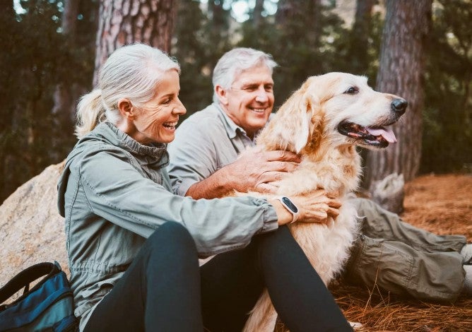 Older man and woman in a wooded area with their golden retriever.