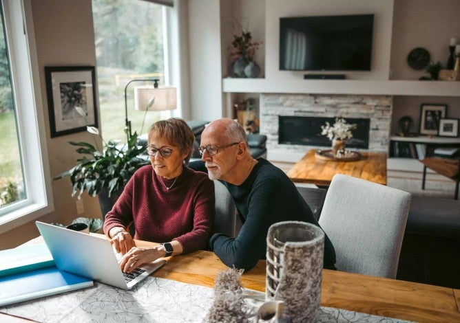 Older couple working together on laptop from dining room table.