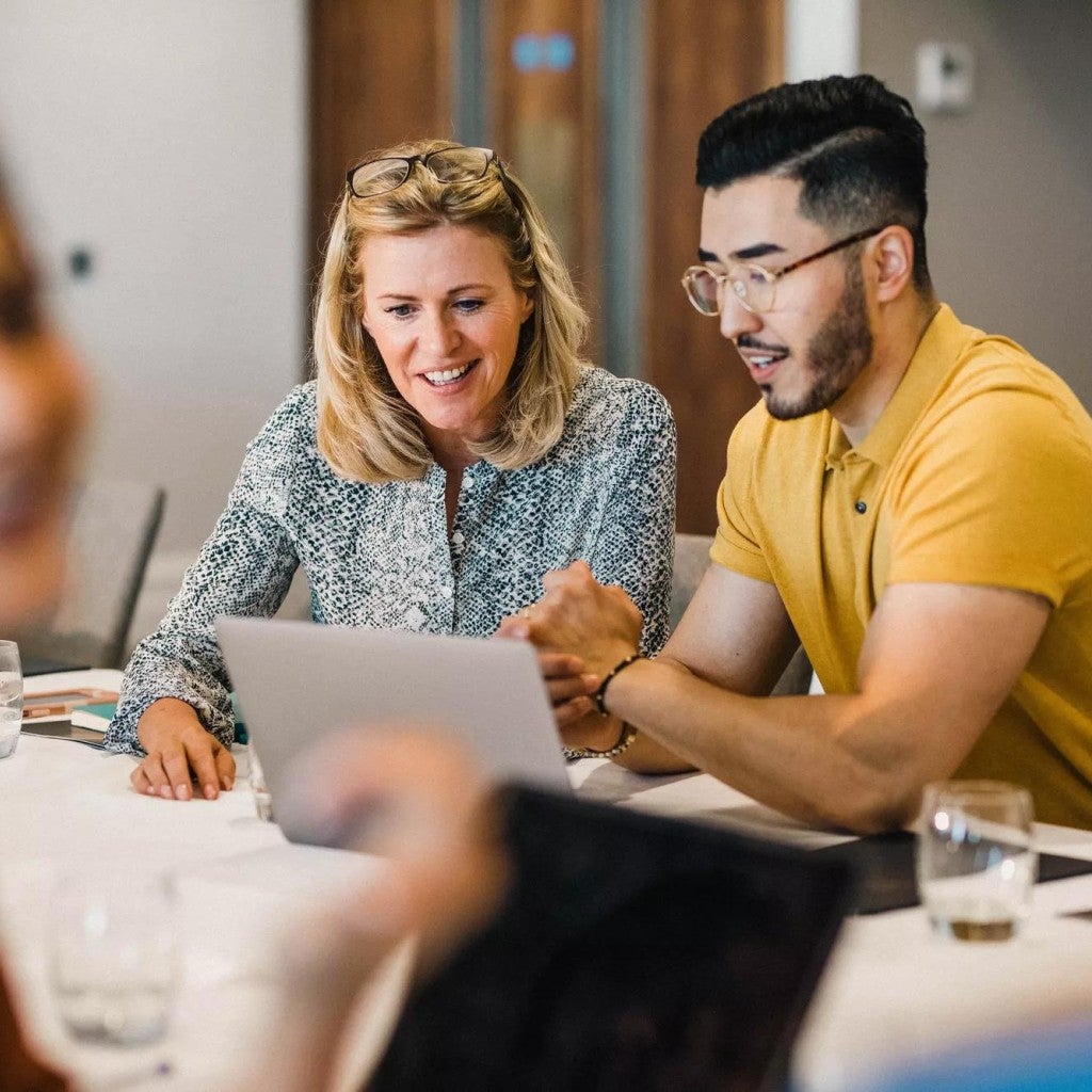Two employees of different generations review a laptop screen at work. Two employees of different generations review a laptop screen at work.