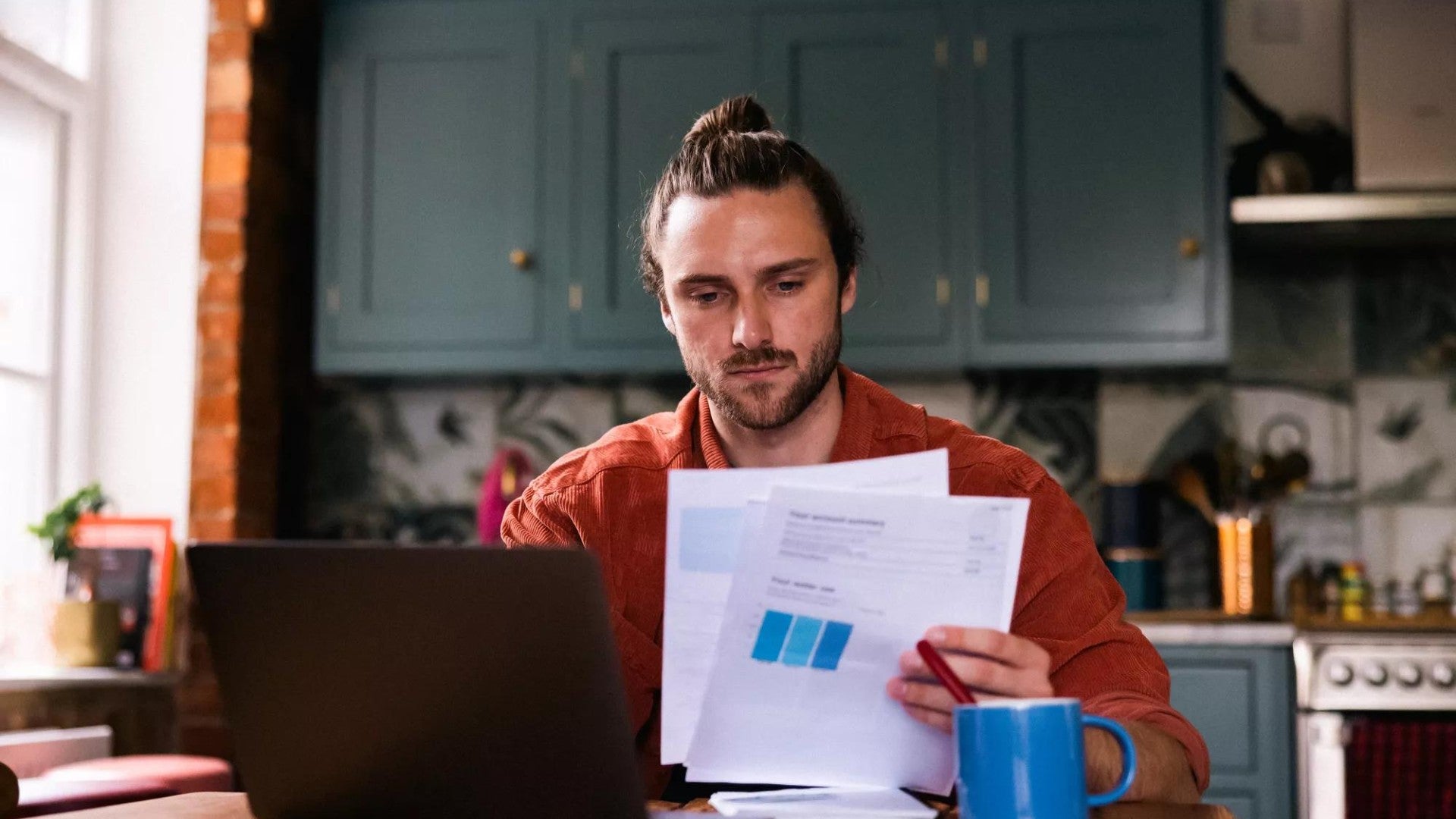 Young adult male at home looking at bills and personal finances using a laptop.