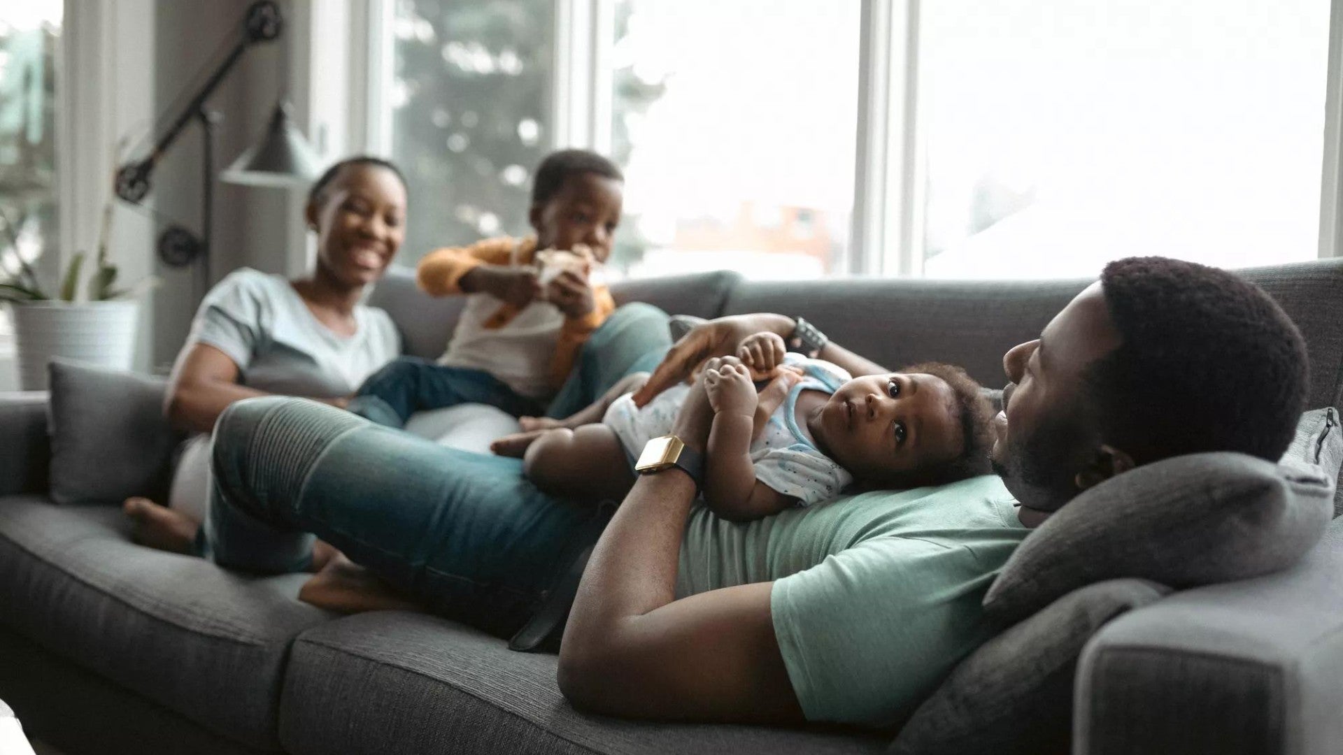 Happy Black family relaxing on a  gray couch with man holding a baby and woman next to a toddler.