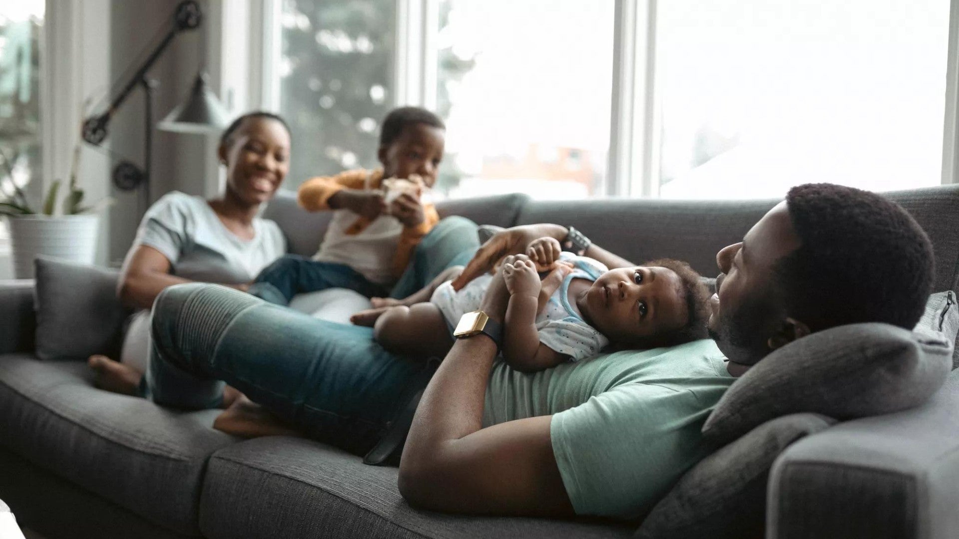 Happy Black family relaxing on a gray couch with man holding a baby and woman next to a toddler.