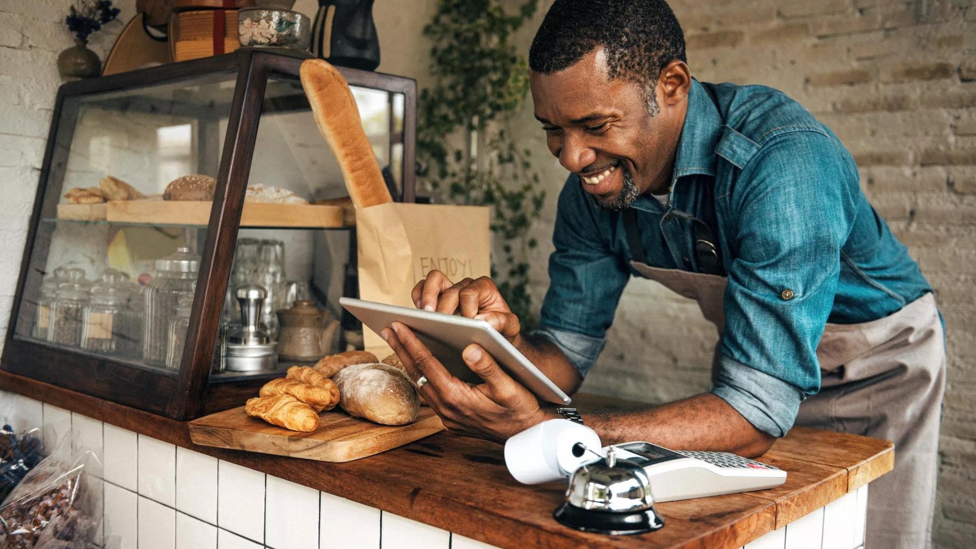 A smiling bakery employee is leaning over a counter and looking at his tablet.