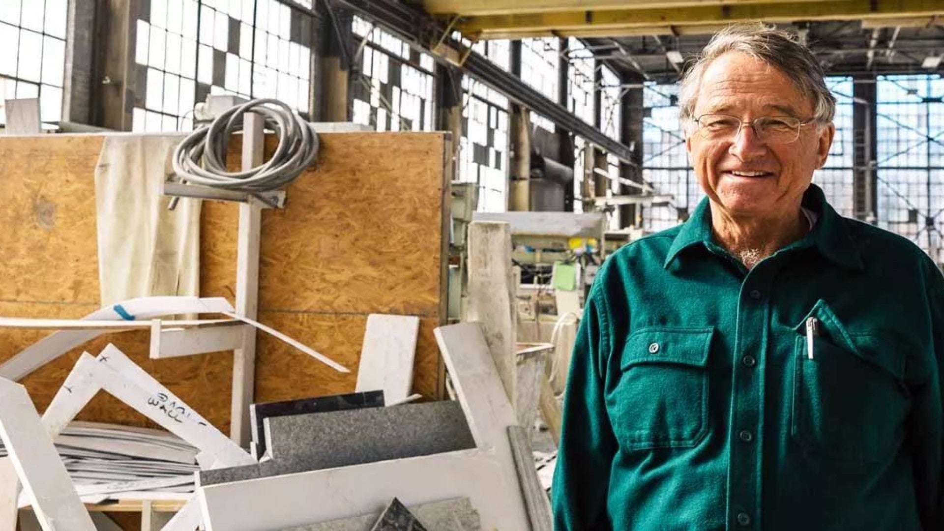 An older entrepreneur stands smiling in a brightly light production facility 