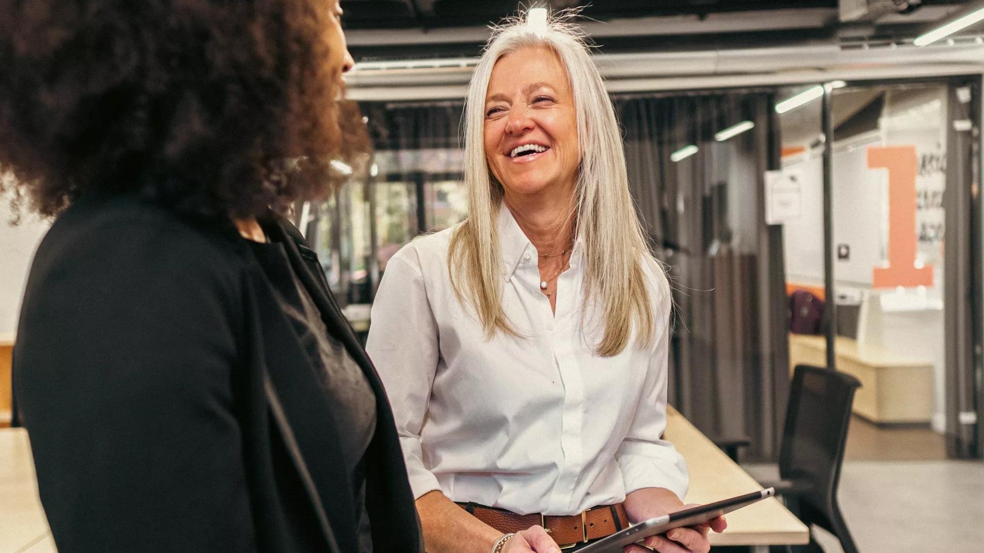 A smiling woman business leader holds a tablet while talking to her female employee in their modern workspace