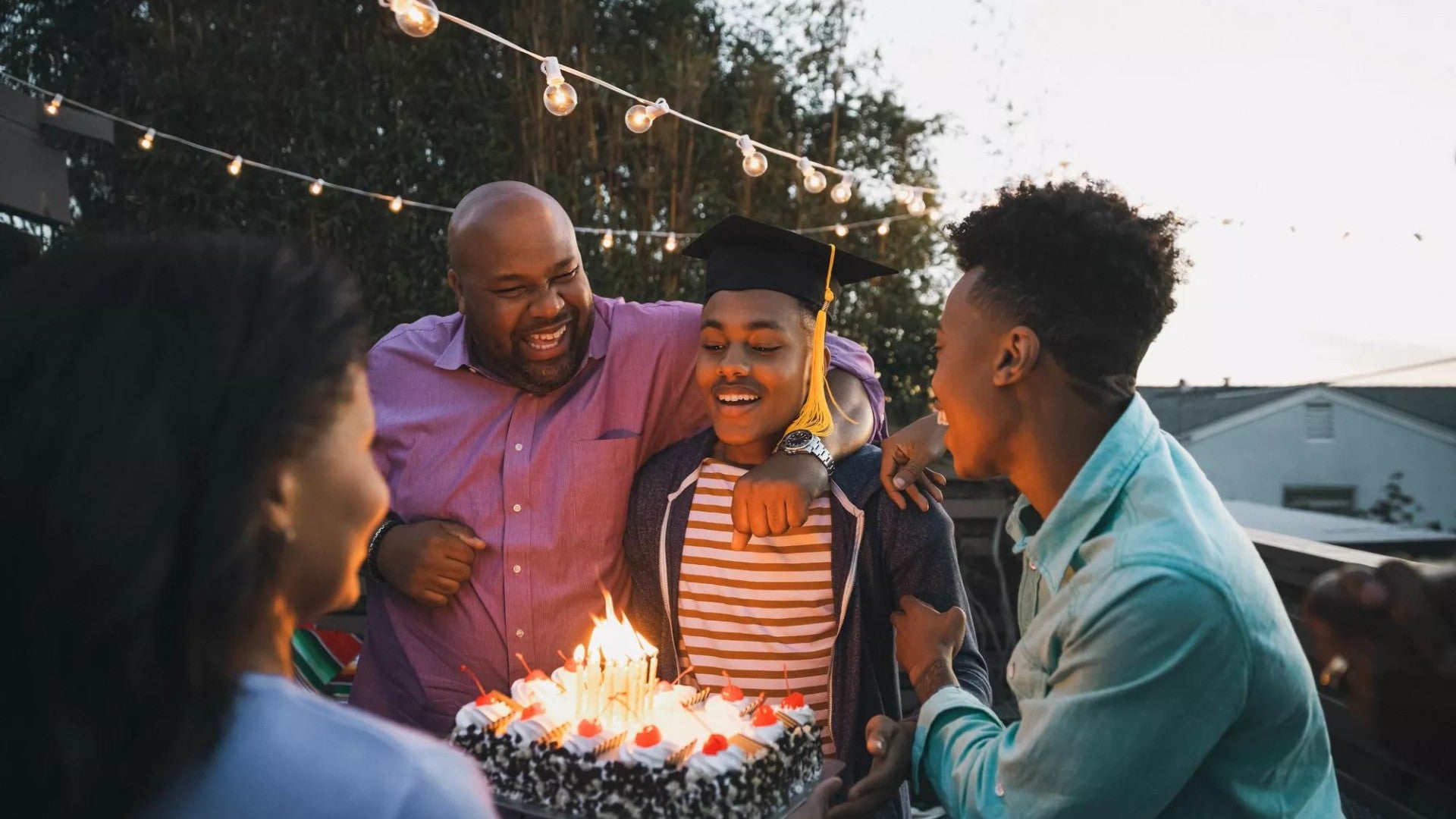 Young man in graduation cap and gown blowing out candles on a cake surrounded by smiling family and friends.