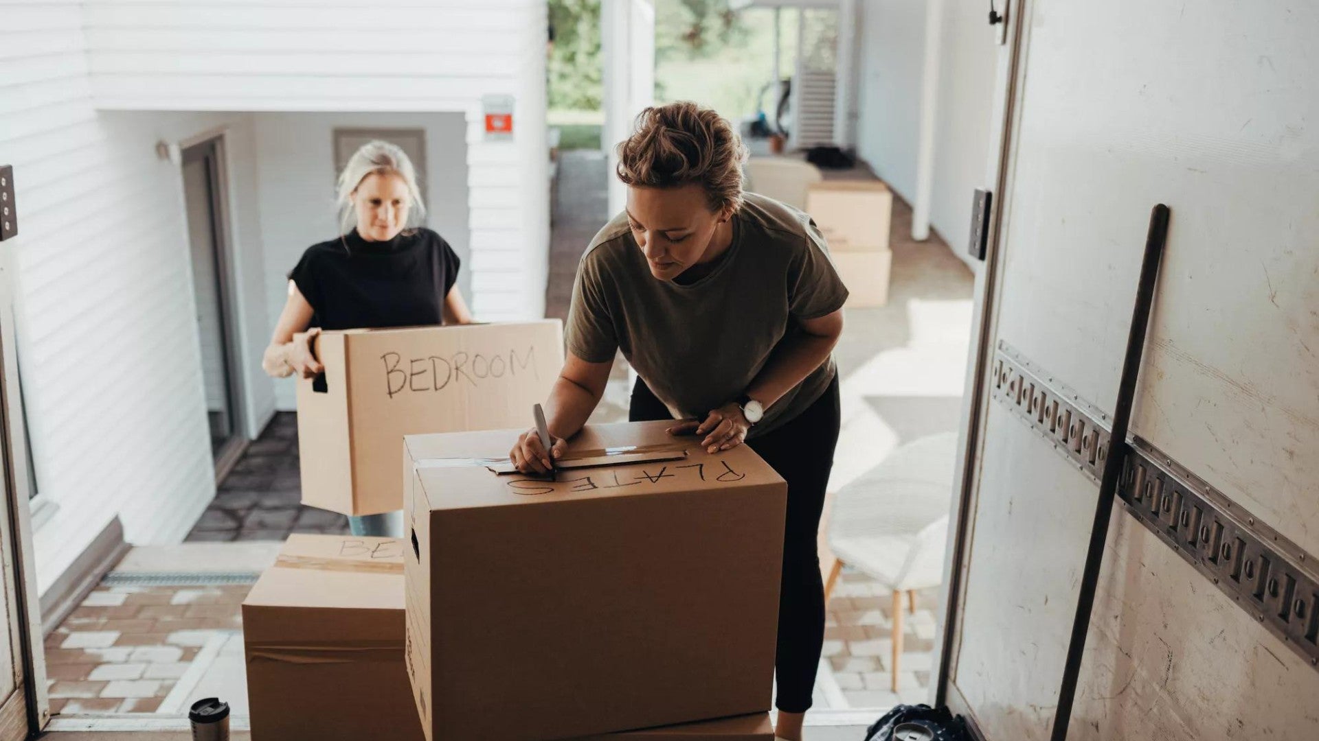 Woman holding box while female writing on cardboard box in moving van