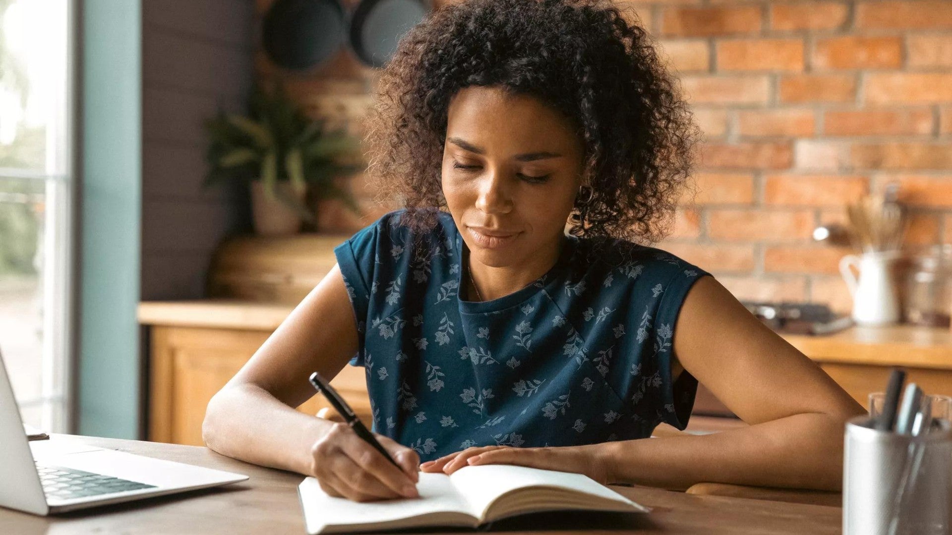 Woman confidently making notes in a notebook while working on her laptop