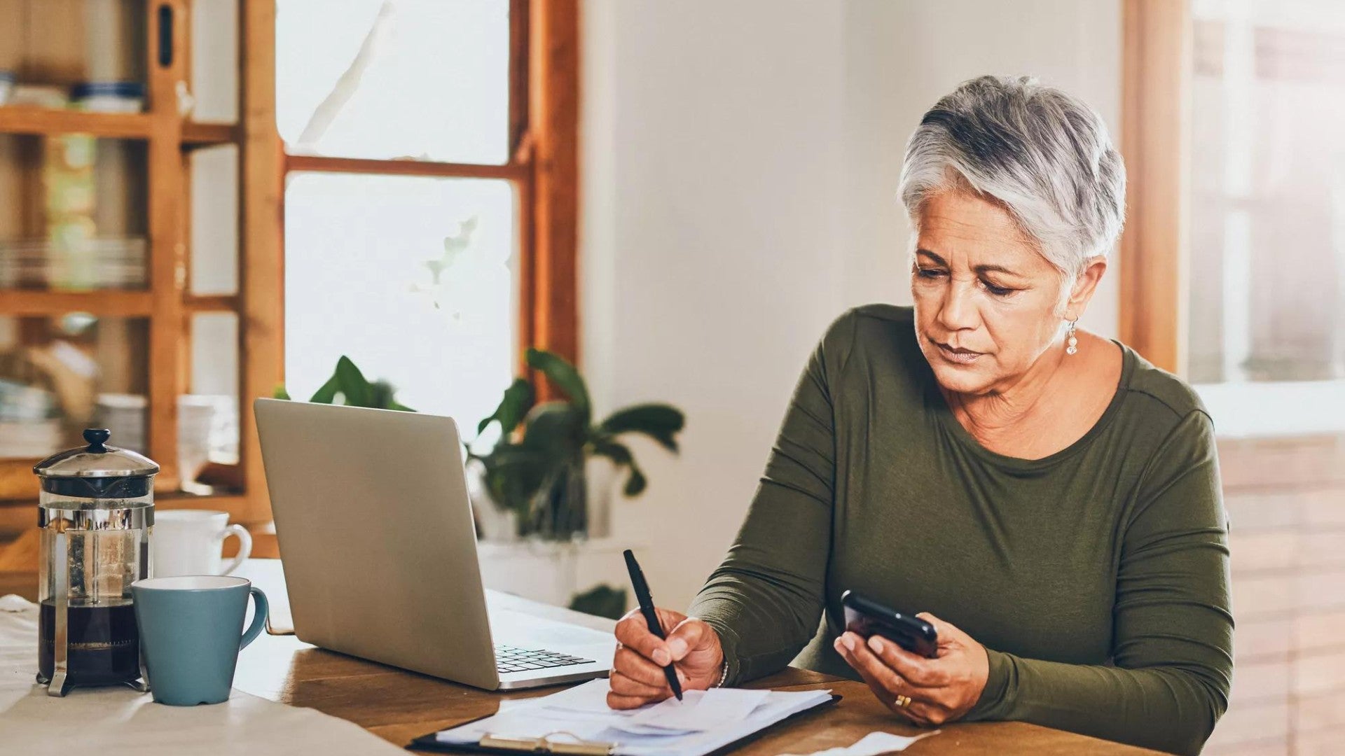 Women working at a table with her laptop and cell phone, writing on paper. 