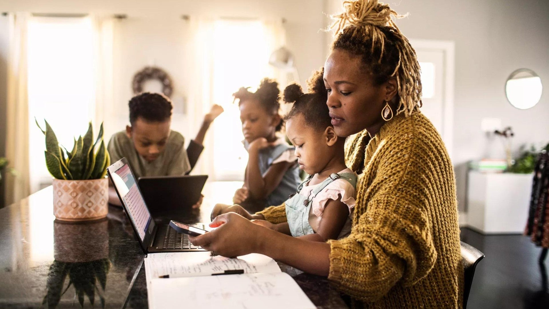 A mother works on her computer at the kitchen counter with her children.