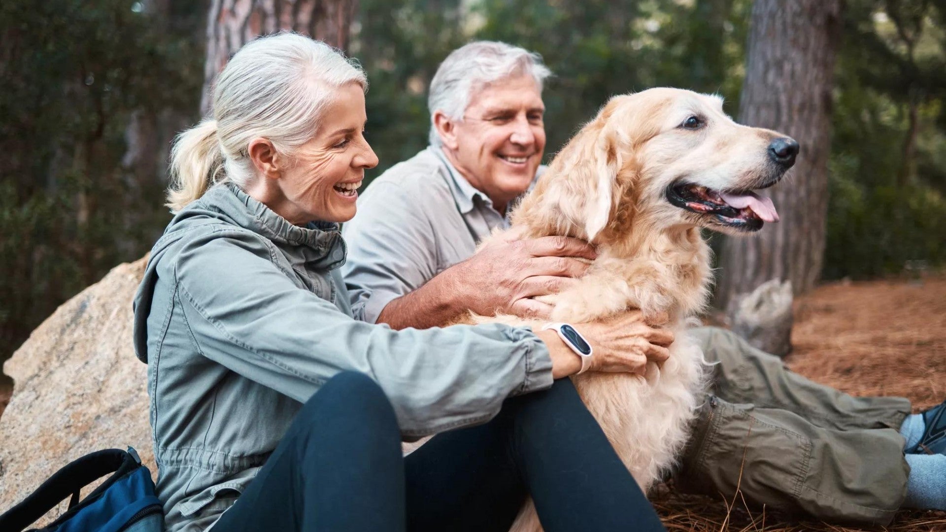 Older man and woman in a wooded area with their golden retriever.