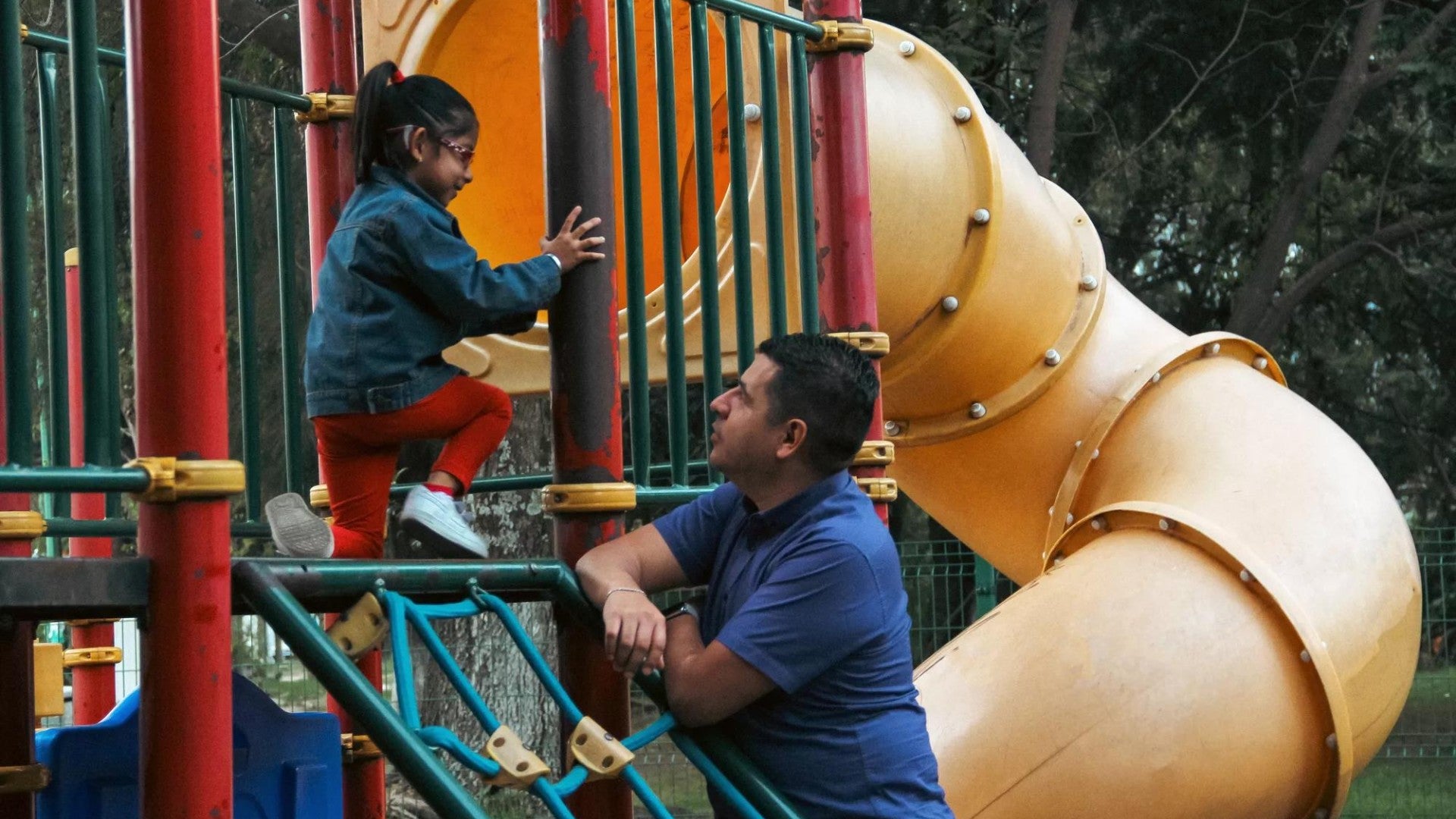 A young girl is on playground equipment that has a big slide; her father is leaning on the equipment looking at her.
